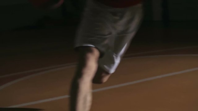 Man dribbling a ball and scoring a point while his opponent trying to prevent him from winning in dark basketball court in slow motion