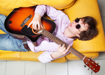 Caucasian male lying on a sofa playing an acoustic guitar and whistles the melody