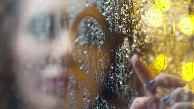 Rack focus from foggy and rainy window pane to smiling woman drawing heart with her finger and looking at camera