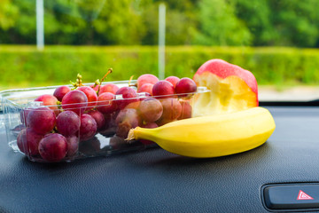 Fruits on dashboard of a car. Grapes in a box, a banana and a half eaten apple lie on black...