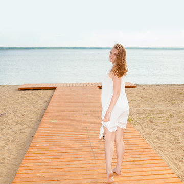 Romantic girl in a white dress with a white hat turned to the long-tailed wooden pier on the beach. The concept of joy and happiness