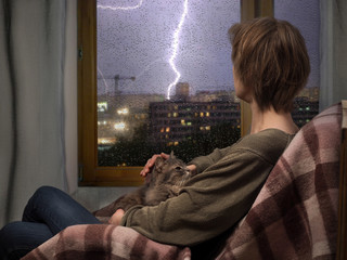 The woman in the room by the window. Night, rain, lightning strikes in the town house. On lap cat. Checkered plaid
