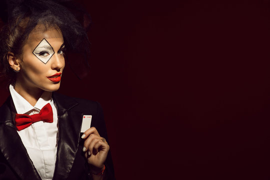 Portrait of a young beautiful lady croupier with an artistic make up joker on the red background taking the ace card out of her tuxedo's lapel. Gamble and casino concept. Studio shot. Copy-space