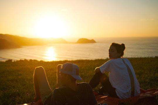 A young surf couple watch sunset over the beach sitting on the grass on the hill