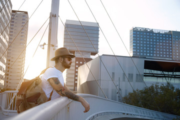 A tattooed and bearded young tourist takes a break at a bridge at sunset against the background of concrete and glass city buildings