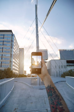 Close up shot of a smartphone with a photo of a city bridge and office buildings held by a tattooed man's arm at sunset