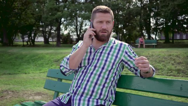 Man talking by smartphone. He is sitting on bench in the park. He is dressed in blue shorts and checkered shirt. He has beard. Steadicam.

