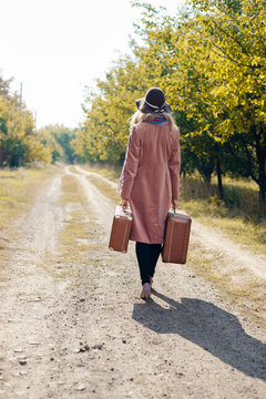 Back view of female wearing hat coat walking away on rural road. She carrying old suitcase over sunny background outdoors