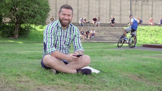 Young man browsing smartphone, tells some words and smiles for camera. He is sitting on grass in the park. He is dressed in blue shorts and checkered shirt. He has beard. Stedicam.
