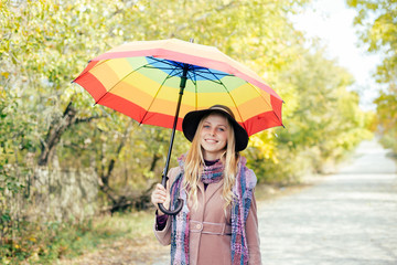 portrait of beautiful young lady with rainbow colorful umbrella having fun happy smiling and looking at camera over autumn road against light sky copy space background