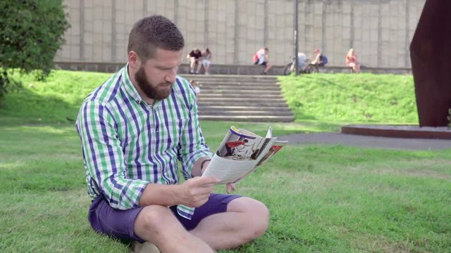 Young man reading magazine and browsing smarphone. He is sitting on grass in the park. He is dressed in blue shorts and checkered shirt. He has beard. Stedicam.
