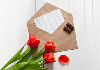 Spring bouquet of red tulips and a card in an envelope on a white wooden background with copy space. View from the top with space for signature.