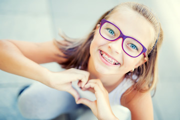 Smiling little girl in with braces and glasses showing heart with hands. © weyo