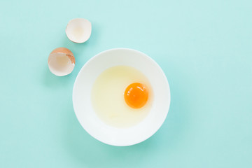 Top view of raw egg in white bowl with eggshell on blue background.