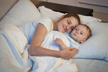 Portrait of happy smiling mother embracing her baby in bed at ni