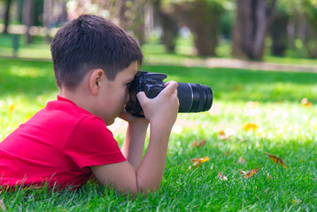 A caucasian boy is interested in the art of photography. A boy in a red t-shirt lies on grass outdoors holding a digital camera, trying to focus on a distant object. Sunny September morning in a park.