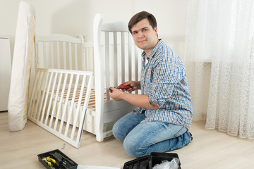 Young man assembling wooden cot in nursery for expectant baby