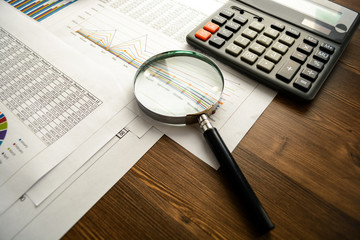 Business accessories (notebook, calculator, planchette, tablet, fountain pen, glasses) and graphics, tables, charts on white sheets on office desk. Soft focus.