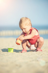 Baby playing on the sandy beach near the sea. Cute little girl in red dress with sand on tropical beach. Ocean coast.
