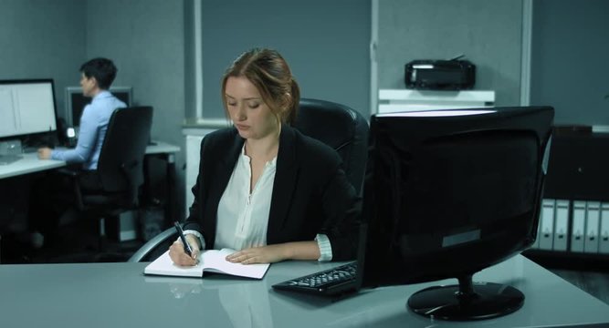 4K: Zwei weibliche Angestellte im B&uuml;ro. W&auml;hrend die Frau im Vordergrund sich Notizen macht, arbeitet die Frau im Hintergrund am PC. Sie unterhalten sich.