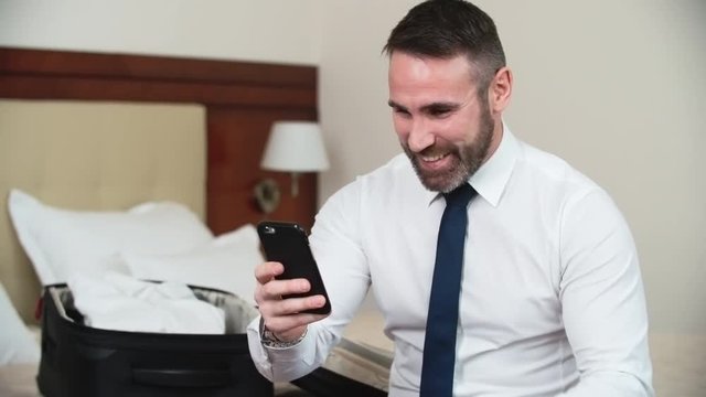 Lockdown shot of middle aged businessman with beard in formal clothes sitting on hotel bed and talking with his family over video call, unpacked suitcase in background 