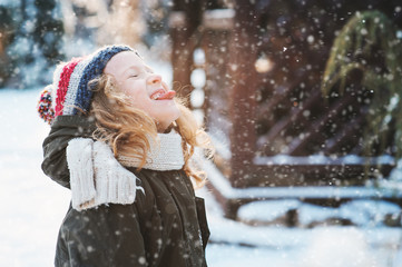 happy child girl catching snowflakes and playing on winter snowy walk in garden, seasonal outdoor...