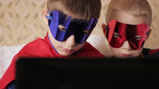 two boys in superhero costumes play a game on the computer, sitting on couch