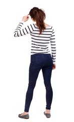 back view of standing young beautiful woman. girl watching. Girl in a striped sweater thoughtfully scratching his head.