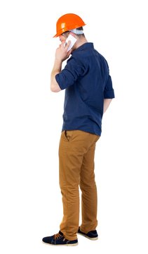 Backview of business man in construction helmet stands and enjoys tablet or using a mobile phone. Isolated over white background. a man in a blue shirt and white helmet talking on a smartphone.