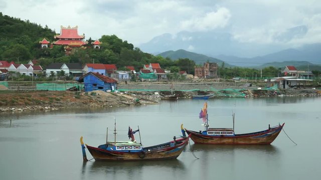 Two fishing boats parked in water near the river bank. On a background there are houses of locals and Buddhist temple into forest.
