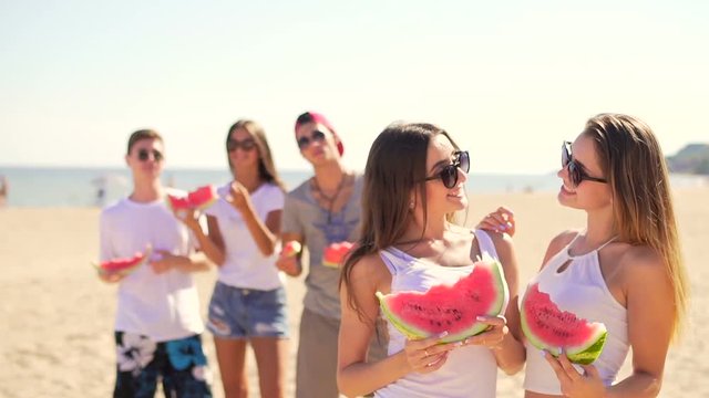 Handsome young women kidding with watermelon at beach. Friends staying at background