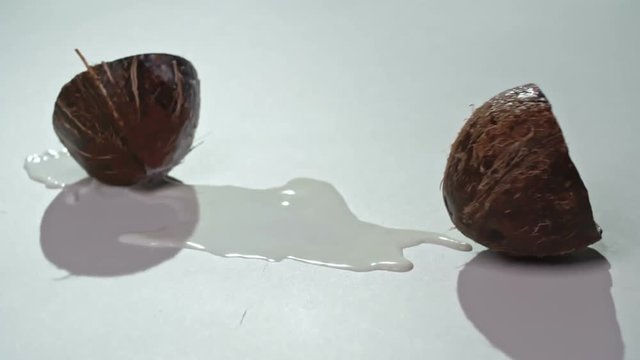 Closeup of coconut falling, splitting into two parts and splashing milk on white surface in slow motion