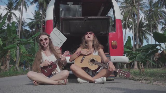 Hippie Girls Play Guitars Sitting on Road near Broken Minivan with an Open Hood. Slow Motion
