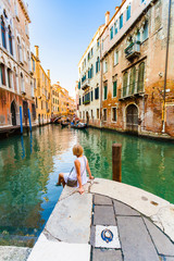 Woman sits near a canal and admires gondolas and architecture of Venice. Italy.