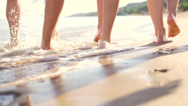 Legs of group of friend while barefoot walking on coast. Sea waves, foams and sprays. Tracking slow motion shot