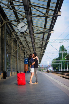 The young pair stand having embraced on platform under clock.