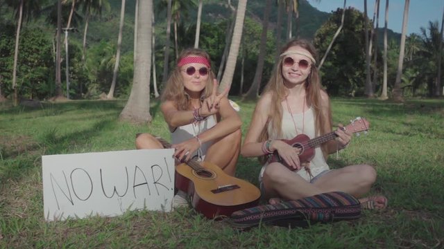 Hippie Girls Play Ukulele Sitting on a Grass in a Palm Grove with an Appeal "No War" on a White Board. Slow Motion