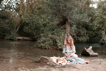 Beautiful young girl having picnic over river. Wearing elegant dress. Posing outdoors. 20s.