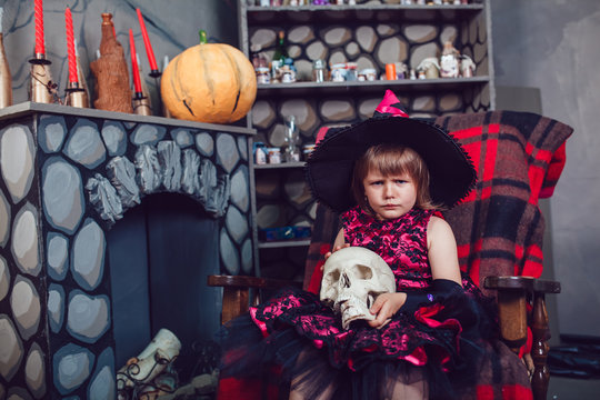 Girl in a witch costume sitting in  chair with  skull in his hands in in halloween decorations