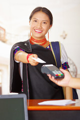Young brunette wearing traditional native clothes working as hotel receptionist with friendly smile, processing payment using credit card terminal, customers point of view