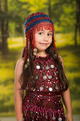 Cute little girl wearing beautiful red dress with matching pearl hat, posing for camera, green forest background