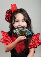 Cute little girl wearing beautiful red and black dress with matching head band, posing for camera using chinese hand fan, studio background