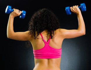 Fototapeta na wymiar Athletic brunette woman wearing pink top, training lifting weights with both arms, back facing camera, black studio background