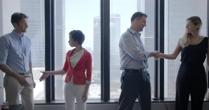 Multi-ethnic team of four business people, men and women, shake hands then hug in front of windows in city office, Downtown Los Angeles. Medium long shot, recorded hand held in slow motion at 60fps