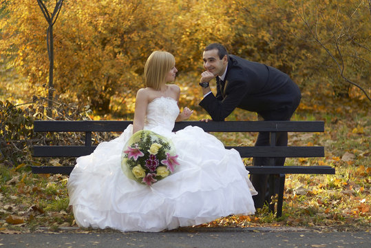 A young couple in love.Bride and groom look at each other with love at picturesque autumn park on the bench.Wedding bouquet,dress/Beautiful married couple in wedding day.Happy smiling newlyweds