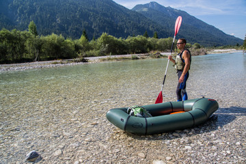 Athletic man in wetsuit standing beside his packraft in a mountain river and looks ahead