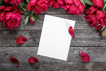 White paper card on rustic wooden background with pink peonies and petals. Flowers. Workspace. Blank for greetings