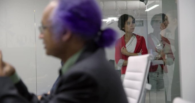 Business woman excluded from meeting, stands outside glass walls of conference room as out-of-focus male colleagues dominate the scene in foreground.   Recorded hand held in real time.