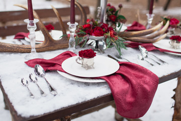 Table setting in red marsala and white colors decorated with candles, napkins, flowers, leaves, horns on wooden table made of pine in winter forest.