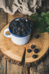 Fresh Blackberries in white cup on round serving wooden board over rustic background, top view, selective focus, vertical composition
